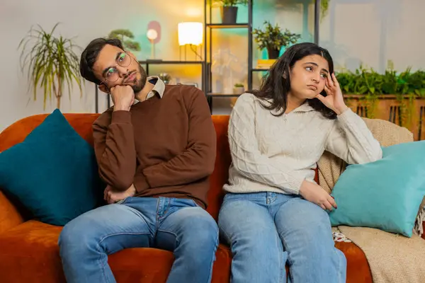 Upset stressed bored tired diverse family couple sitting on sofa at home room apartment. Man with Indian girlfriend. Trusting relations compassion. Problem, depressed feeling bad annoyed, burnout