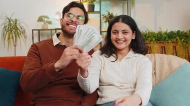 Mutlu genç Hintli çift ellerinde para banknotları tutarken odadaki kanepede oturan kameraya bakıyorlar. Başarılı zengin ve çeşitli aile yumrukları piyango ikramiyesini kazandı.