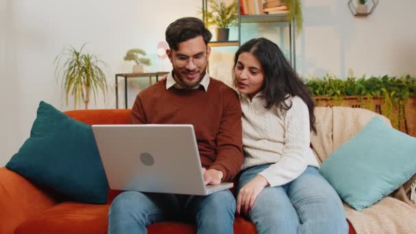 在家里客厅的沙发上使用笔记本电脑时 穿着休闲装的快乐而又多姿多彩的年轻夫妇在聊天 快乐的计划生育一起指向网上订票的公寓 — 图库视频影像