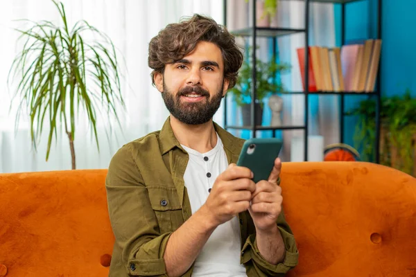 Middle eastern man sits on couch uses mobile phone smiles at home living room apartment. Young guy texting share messages content on smartphone social media applications online, watching relax movie