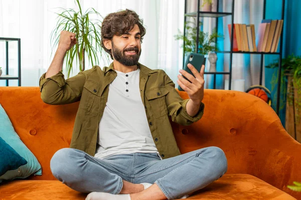 Happy man use mobile smartphone typing browsing say Wow yes, celebrating success victory, winning lottery jackpot goal achievement, play game, good positive news, triumph. Guy at home in room on sofa