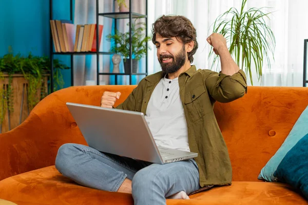 Surprised middle eastern man using laptop computer, receive mail good news message shocked by sudden victory celebrate lottery jackpot win purchases online shopping play game at home apartment on sofa