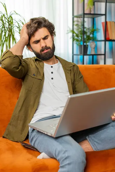 Displeased Middle eastern man use laptop notebook typing browsing working, loses becoming surprised sudden lottery results bad news fortune loss game fail computer virus. Young guy on home room couch