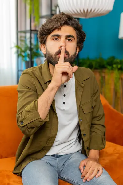 Shh be quiet please. Middle eastern man presses index finger to lips makes silence hush gesture sign do not tells gossip secret. Arabian guy at home apartment living room sitting on couch. Vertical