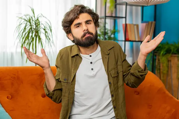 I dont know what to say. Confused middle eastern Arabian man feeling embarrassed about ambiguous question, having doubts, no idea, being clueless and uncertain. Young bearded guy at home room on couch
