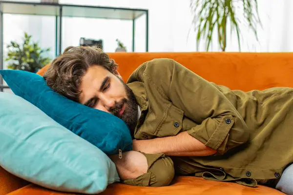 Tired middle eastern arabian man lying down in bed taking a rest at home. Carefree young bearded guy napping, falling asleep on comfortable sofa couch with pillows. Closed eyes enjoy daytime nap alone