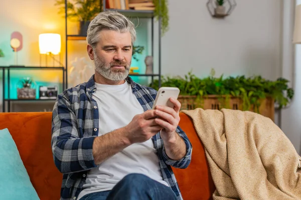 Happy mature man sitting on sofa at home getting message from old friend using smartphone look aside with smile. Caucasian grey-haired old man reading good news on phone screen in living room at home
