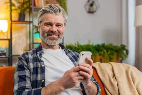 Happy mature man sitting on sofa at home getting message from old friend using smartphone look aside with smile. Caucasian smiling old man reading good news on phone screen in living room at home