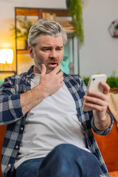 Shocked sad mature man holding smartphone reading negative message feels annoyed sitting on sofa in living room. Senior bearded old guy having gadget trouble in apartment. App crash concept. Vertical
