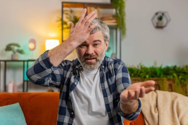 Webcam görüntüsü. Memnuniyetsiz, orta yaşlı, sakallı, tartışan, kavgacı, video görüşmesinde evdeki kanepede oturan adam. Kızgın, olgun bir erkek, kameraya bakmaktan şikayetçi.