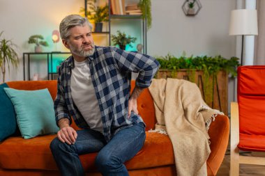 Oturma odasındaki kanepede oturmaktan sırtı ağrıyan yaşlı ve yorgun bir adam. Üzgün yaşlı erkek masajı ağrılı bel kasları. Kafkasyalı, olgun, yorgun, çok çalışan bir adam..