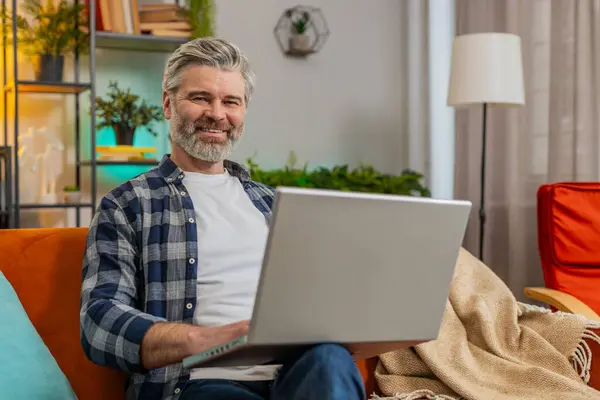 Smiling bearded mature man typing on laptop browsing internet using apps sitting on sofa alone. Happy middle-aged senior Caucasian old guy working on computer looking at camera in living room at home