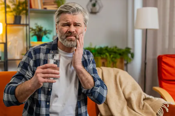 Senior man feel terrible toothache after drink cold water. Middle-aged guy sits on sofa in living room touching cheek, feel hurt and suffering from sensitive tooth ache, pain and cavities. Copy-space