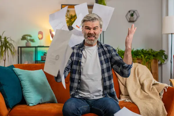 Dissatisfied mature man throwing out bills, checks, or papers and documents while sitting on couch at home. Middle aged Caucasian old guy gets rid of paperwork accounting looking away at living room.