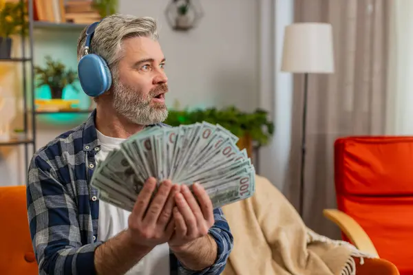 Happy excited rich joyful senior man in wireless headphones showing wasting money cash around, more tips earnings, big profit, win lottery jackpot share celebrating success on sofa at home. Copy-space