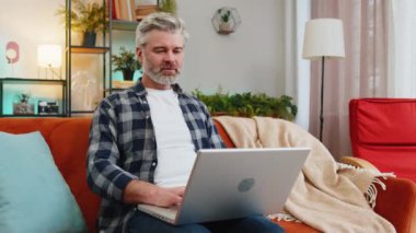 Gülümseyen sakallı, olgun bir adam dizüstü bilgisayarda yazı yazıyor. İnternette geziniyor. Kanepede tek başına oturan uygulamaları kullanıyor. Orta yaşlı, yaşlı, beyaz bir adam bilgisayarda çalışıyor. Oturma odasında ekrana bakıyor.