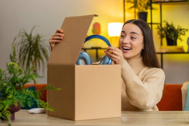 Evde oturan mutlu beyaz kadın teslimat paketini açıyor. Gülümseyen kız alışverişci, çevrimiçi dükkan müşterisi karton kutu açıyor posta yoluyla hediye kulaklıkları alıyor