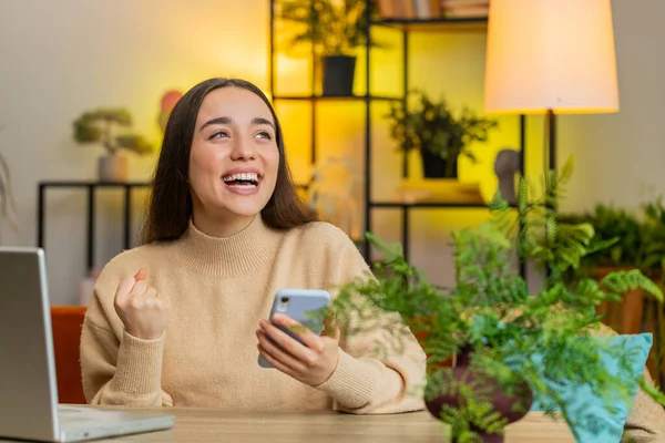 Happy woman use mobile smartphone browsing say Wow yes celebrating success victory winning lottery jackpot goal achievement play game good positive news triumph. Girl at home room table. Copy-space