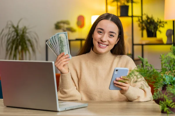 Planning budget. Rich happy Caucasian young woman counting money cash use laptop computer calculate domestic bills. Female girl satisfied of income earnings saves money sitting at home office at table