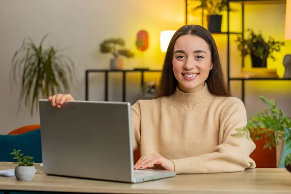 Happy smiling Caucasian young woman freelancer opens laptop working on notebook, sends messages business email makes online purchases watching movies. Female girl sitting at home office room at table