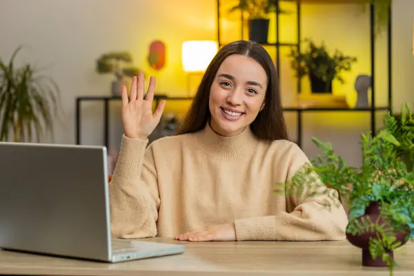 Hello. Pretty young woman smiling friendly at camera, waving hands gesturing hello hi, greeting or goodbye welcoming with hospitable expression at home office. Caucasian girl sitting at table in room
