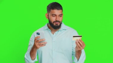 Kredi kartı ve akıllı telefon kullanan mutlu genç Hintli adam harcamaları transfer ederken, online alışveriş yaparken, yemek siparişi verirken. Yeşil krom anahtar arka planında izole edilmiş bir Arap.