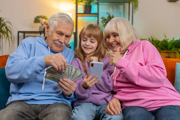 Caucasian grandfather, grandmother, granddaughter counting money using smartphone calculator at home. Smiling excited girl and senior grandparents celebrating success winning lottery sitting on sofa
