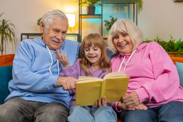 Caucasian grandparents and little granddaughter reading interesting book together sitting on sofa in living room at home. Girl with grandparents spending leisure time on comfortable couch in apartment