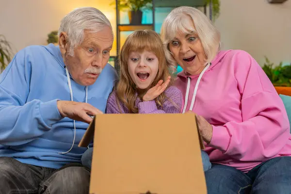 Happy excited Caucasian grandparents and granddaughter girl opening cardboard box together sits on sofa at home. Shocked family consumers unpack good parcel looking inside great purchase delivered.