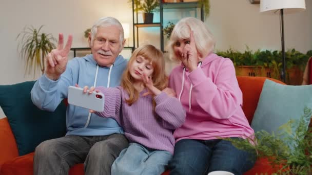 高兴的祖父 祖母和孙女在家里用智能手机自拍自拍 与祖父母一起笑着的白人女孩让博客写手坐在沙发上展示和平标志 — 图库视频影像