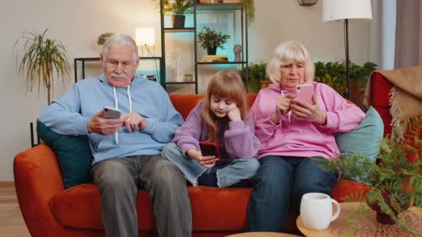 高加索人的祖父 祖母和孙女在家里客厅里使用智能手机 微笑的女孩和老夫妇在沙发上度过闲暇时光 玩网络游戏 社交媒体 — 图库视频影像