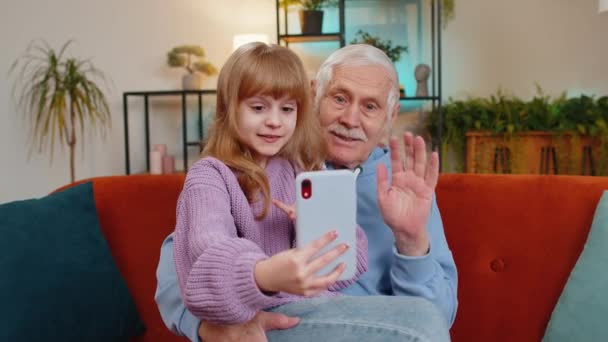 白人の祖父 かわいい孫娘が自宅でスマートフォンでオンラインでビデオ通話をする インターネットウェブカムチャットアプリで話す祖父母を持つ幸せな少女 ソファーで挨拶 — ストック動画