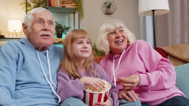快乐的白种人祖父 祖母和孙女在家里吃爆米花 在沙发上看电影 周末和祖父母一起在客厅里看电影的小女孩笑着 — 图库视频影像