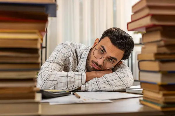 Exhausted Arabian businessman reviewing documents reports while using calculator working hard in home office. Stressed tired freelancer touching head in despair surrounded by stacks of books on table