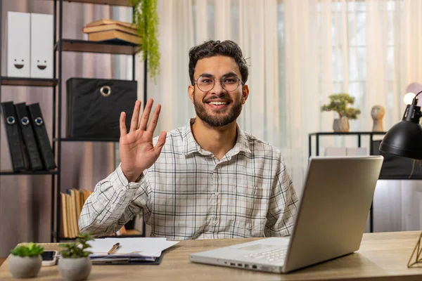 Hello. Happy young Indian businessman employee waves hand hi gesture greeting welcomes someone, online webinar, remote distance conversation. Freelancer guy sitting at desk with laptop in home office.