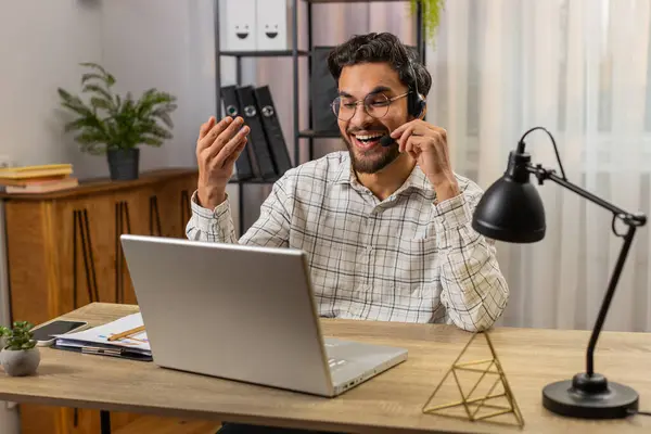 Young Indian businessman wearing headset, freelance worker, call center or support service operator helpline, talking with client or colleague communication support. Guy sitting at home office desk.