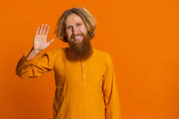 Young Caucasian man smiling friendly at camera, waving hands gesturing hello greeting or goodbye welcoming with invitation hospitable expression. Bearded guy isolated on orange studio background