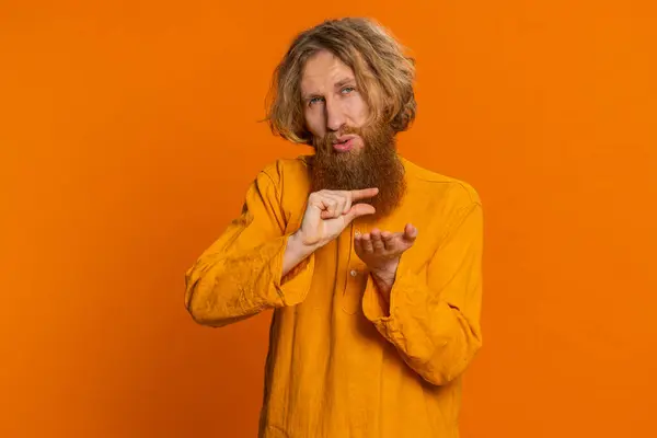 Need some more, please give me. Caucasian man showing a little bit gesture with sceptic smile showing minimum sign, measuring small size begging help. Redhead guy isolated on orange studio background