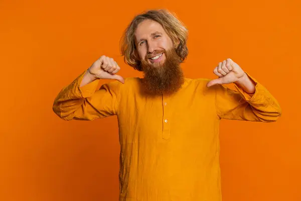 I am the best, choose me. Happy Caucasian man feeling very proud pointing himself, looking self-confident, overjoyed by success, making choice. Young guy isolated on studio orange background indoors