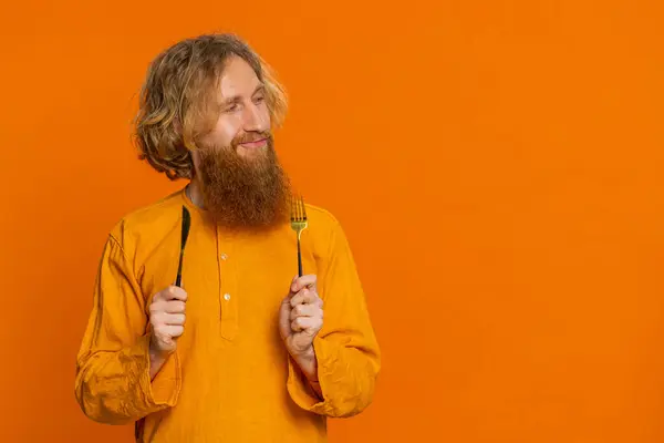 Ready to eat. Hungry Caucasian man waiting for serving dinner dishes with with restlessness holding cutlery fork knife, will appreciate delicious restaurant meal. Redhead guy on orange background