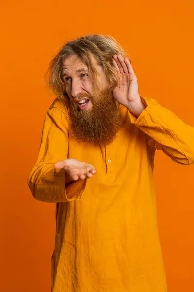 I cant hear you. What. Caucasian man trying hear you looking confused and frowning keeping arm near ear for louder voice asking to repeat to hear information deafness. Bearded guy on orange background