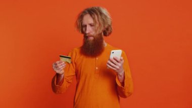 Kredi kartı ve akıllı telefon kullanan Kafkasyalı mutlu bir adam harcama parasını transfer ederken, online alışveriş yaparken, yemek siparişi verirken. Kızıl saçlı genç adam turuncu arka planda kapalı alanda izole edilmiş.
