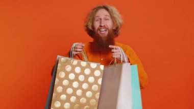 Alışveriş torbaları, reklam indirimleri, düşük fiyatlarla şaşkın şaşkın bakan, Kara Cuma tatillerinde alışveriş yapan mutlu beyaz adam. Kızıl saçlı genç adam turuncu arka planda izole edilmiş. Kopyalama alanı