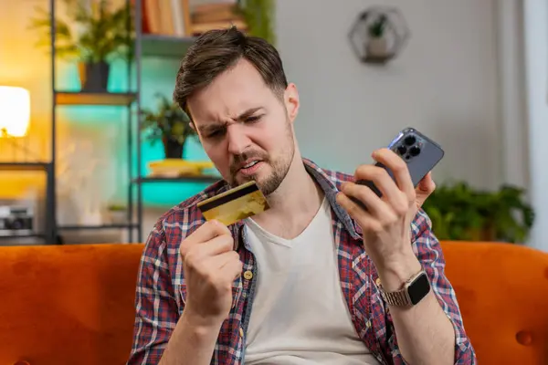 Sad young man guy in plaid shirt sitting on sofa holding smartphone and credit card frowns trying to make payment through internet feels angry due lack of money. Caucasian victim of fraud concept