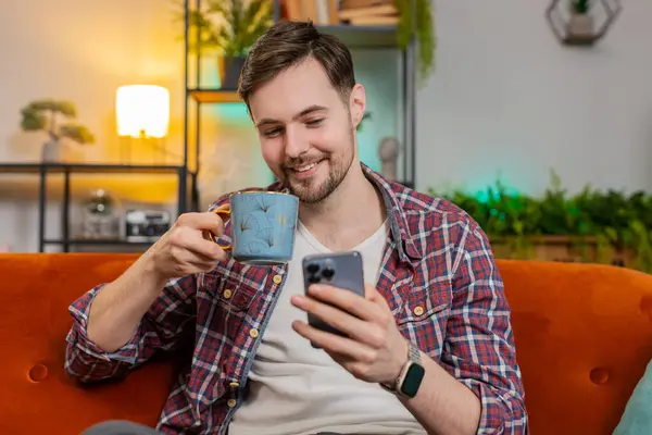 Happy young man in casual shirt uses smartphone while having hot coffee from cup. Caucasian bearded guy having chat on social media using cellphone enjoying chatting to friend sitting on sofa at home.