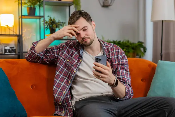 Shocked sad young man using smartphone reading negative e-mail message feels annoyed sitting on sofa in living room. Caucasian guy having gadget trouble problem in apartment. App virus crash concept.