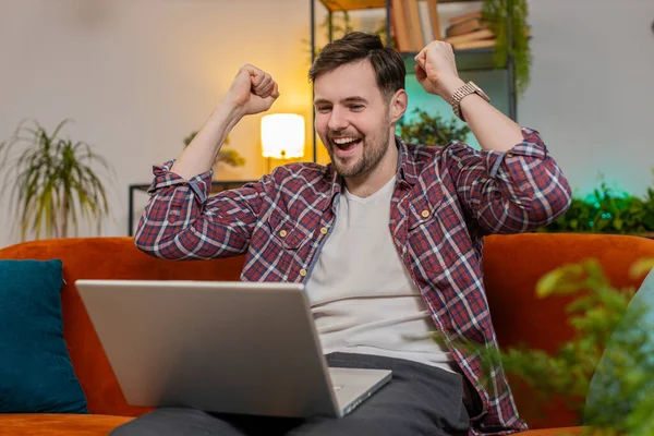 Surprised amazed man using laptop receive good news shocked by sudden victory celebrate lottery jackpot win in apartment. Cheerful Caucasian guy with laptop sitting on sofa in living room at home.