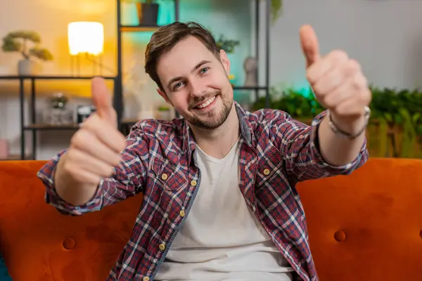 Portrait of excited Caucasian man looking approvingly at camera showing double thumbs up, like sign satisfied, good news, positive feedback. Young happy guy sitting on couch in living room at home