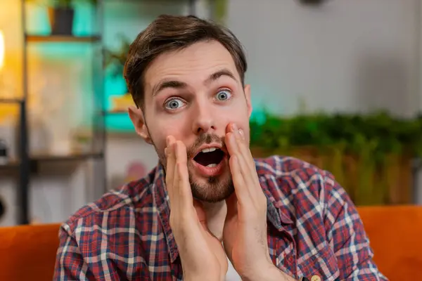 Portrait of amazed shocked man in plaid shirt with wow reaction open mouth looking at camera in living room at home. Happy excited Caucasian guy with big eyes shocked by sudden victory good win news