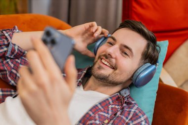 Kablosuz kulaklıklı mutlu beyaz adam akıllı telefondan en sevilen enerjik dans müziğini dinliyor. Hafta sonları evdeki oturma odasındaki kanepede dinlenen gülümseyen adam.